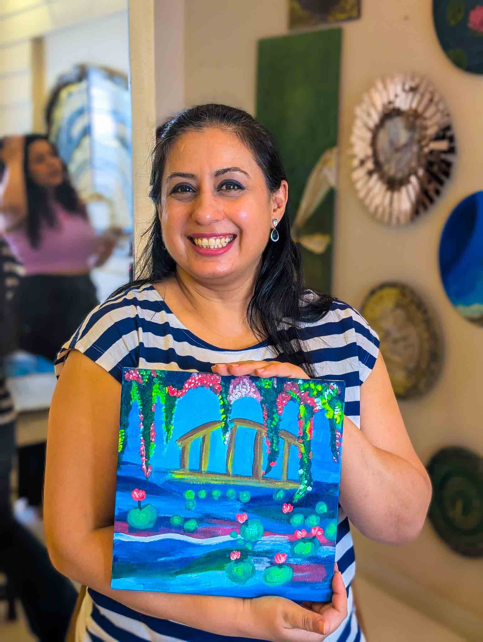Smiling woman showing her completed bright blue painting of a bridge and water lilies in a beginner-friendly Sip & Paint class.